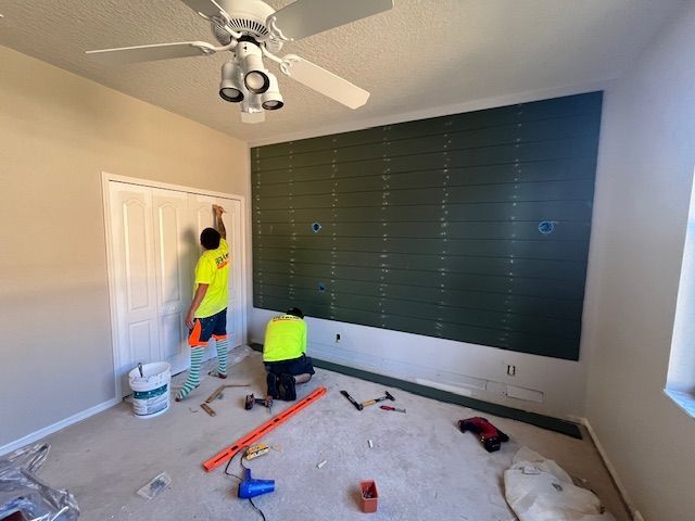 Two workers install green paneling on a bedroom wall. One stands, reaching up; the other is crouched down.