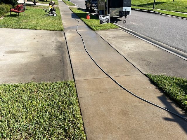 Sidewalk being power washed; a dark hose snakes across the concrete. Green grass surrounds the walkway.