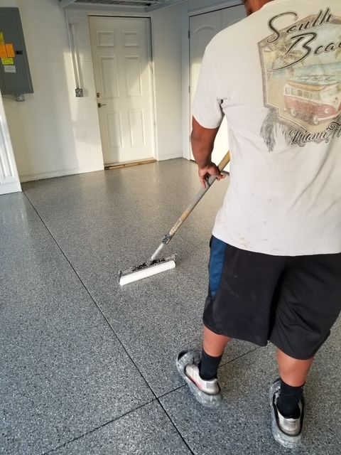 A person applies coating to a gray epoxy garage floor. They use a roller on an extended handle.