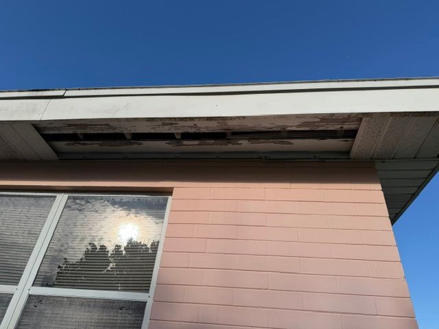 House eaves with damaged soffit above a window, against a clear blue sky