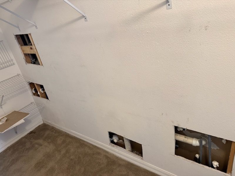 Interior wall with multiple rectangular holes revealing pipes, wiring, and framing. Beige carpet.