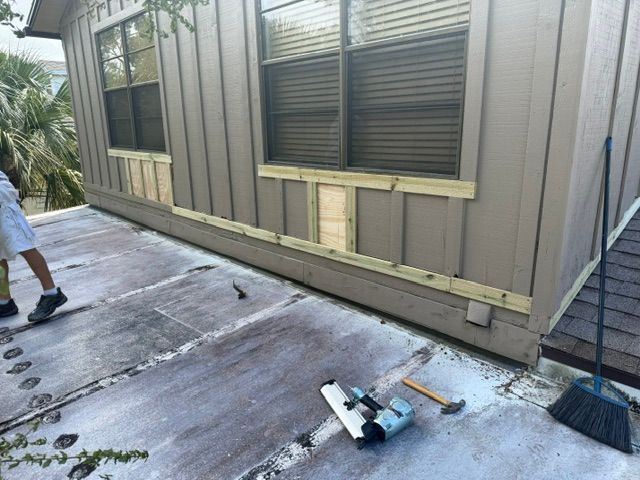 House exterior with damaged lower siding, a person at left, and tools on the driveway near a broom.