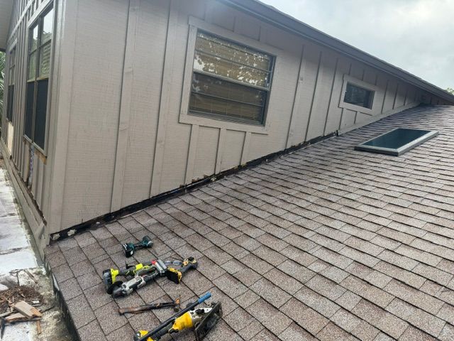 Roofing tools on a shingled sloped roof beside a house wall and window.