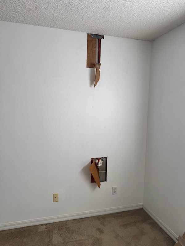 White wall with two holes exposing wooden structures, carpeted floor, light switch.