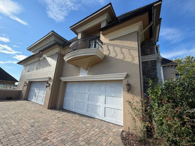 Two-story beige house with white garage doors and a small balcony over a brick driveway