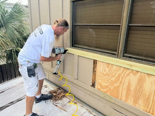 Person using a drill on plywood covering a wall beneath windows outdoors