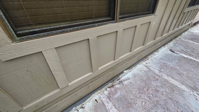 Tan house exterior wall beneath a window, beside a concrete walkway with snow patches