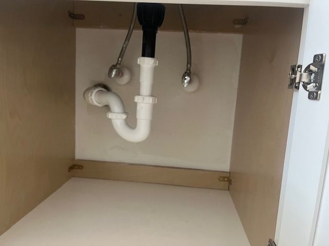 Inside of a bathroom cabinet with plumbing pipes. White P-trap, chrome and black pipes on white wall.