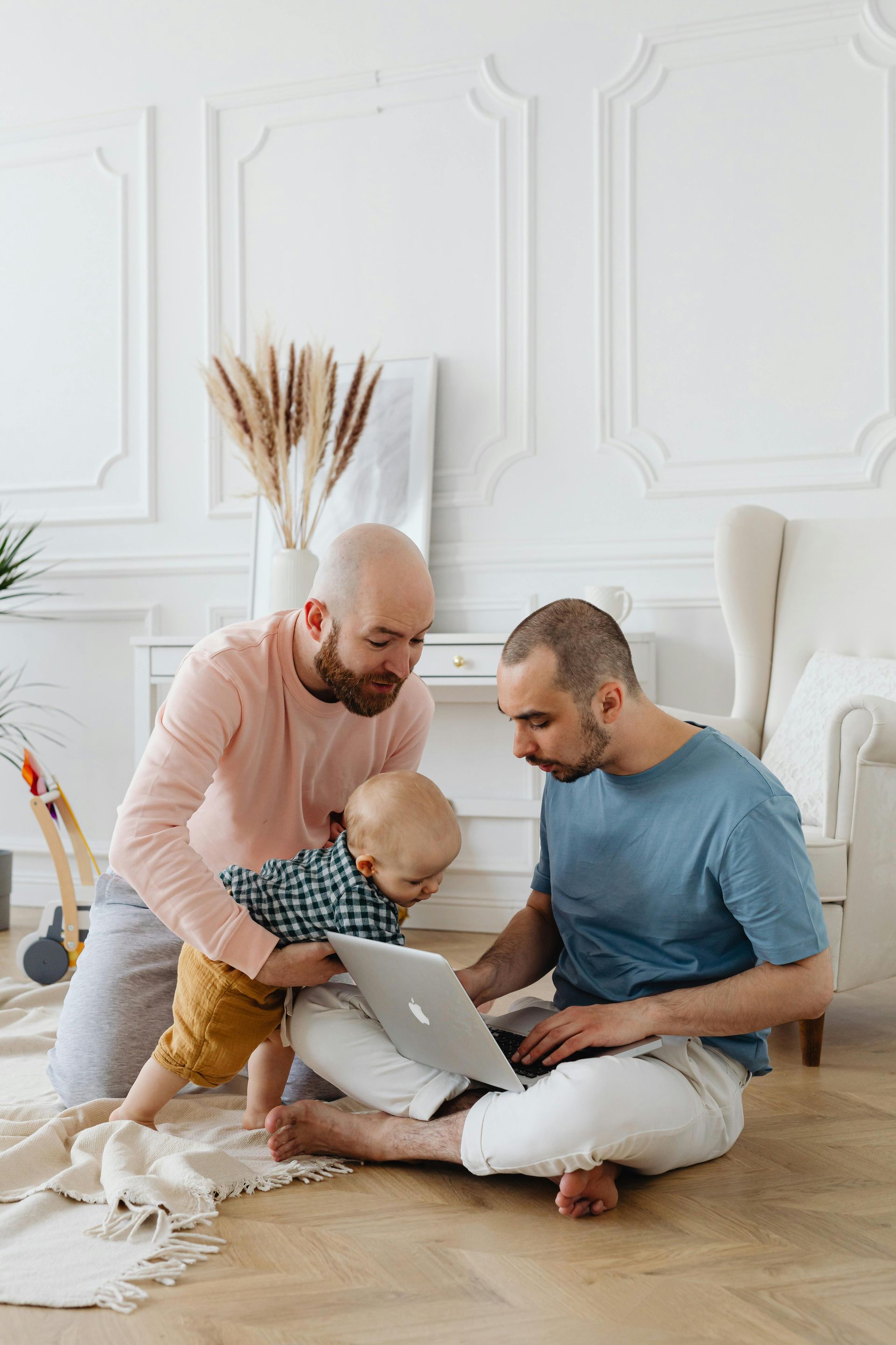 Two men are sitting on the floor with a baby and a laptop.