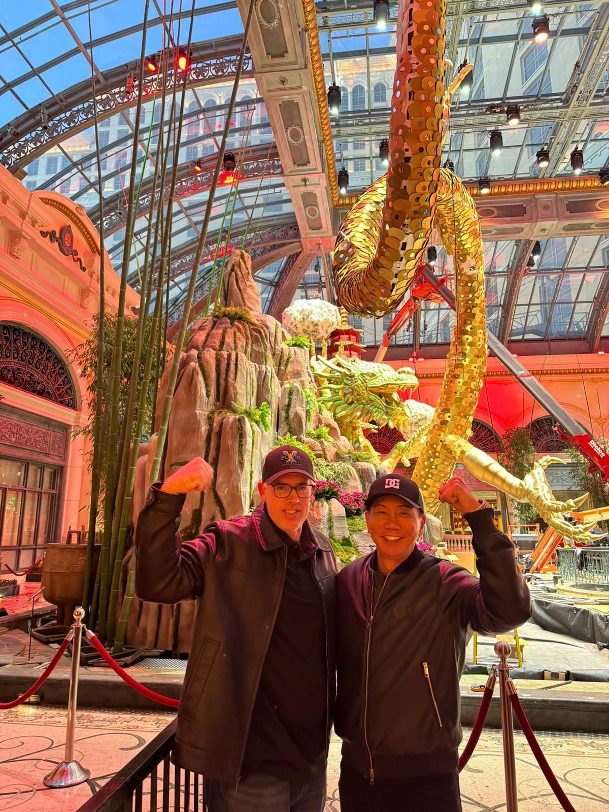 Two people posing beneath a giant golden dragon sculpture in an indoor glass-roofed atrium