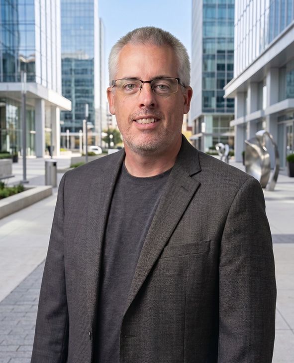 Man in a gray blazer standing outdoors in a modern office plaza with glass buildings behind him
