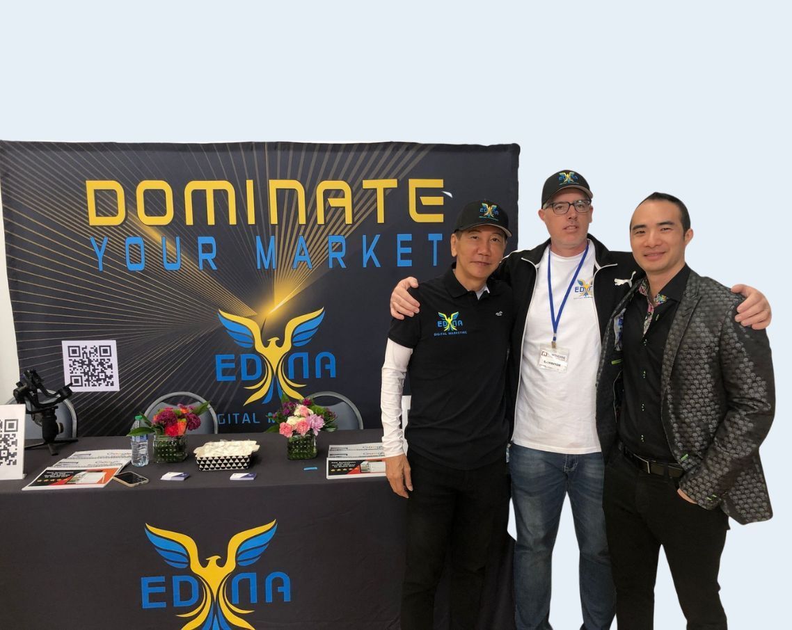 Three people pose beside a Domin8 table display with black and yellow branding and sunglasses on the table.