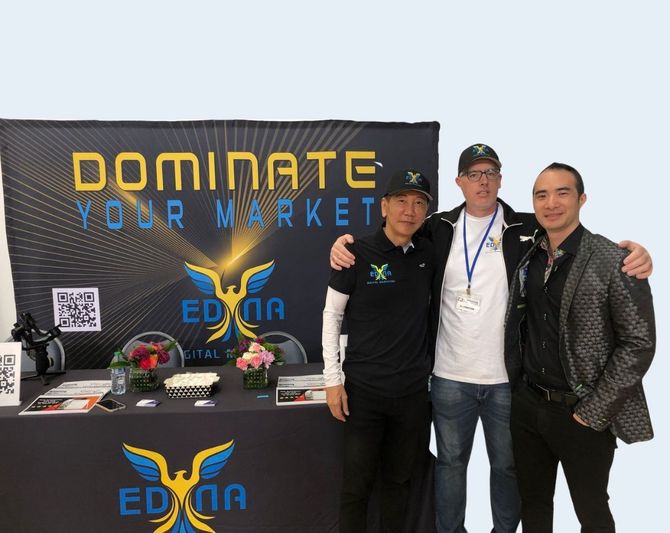 Three people pose beside a Domin8 table display with black and yellow branding and sunglasses on the table.