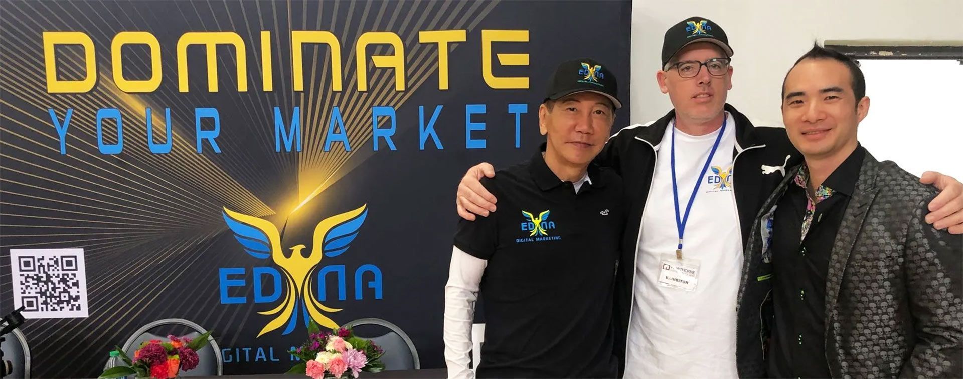 Three men posing at a market-themed event backdrop reading “Dominate Your Market.”