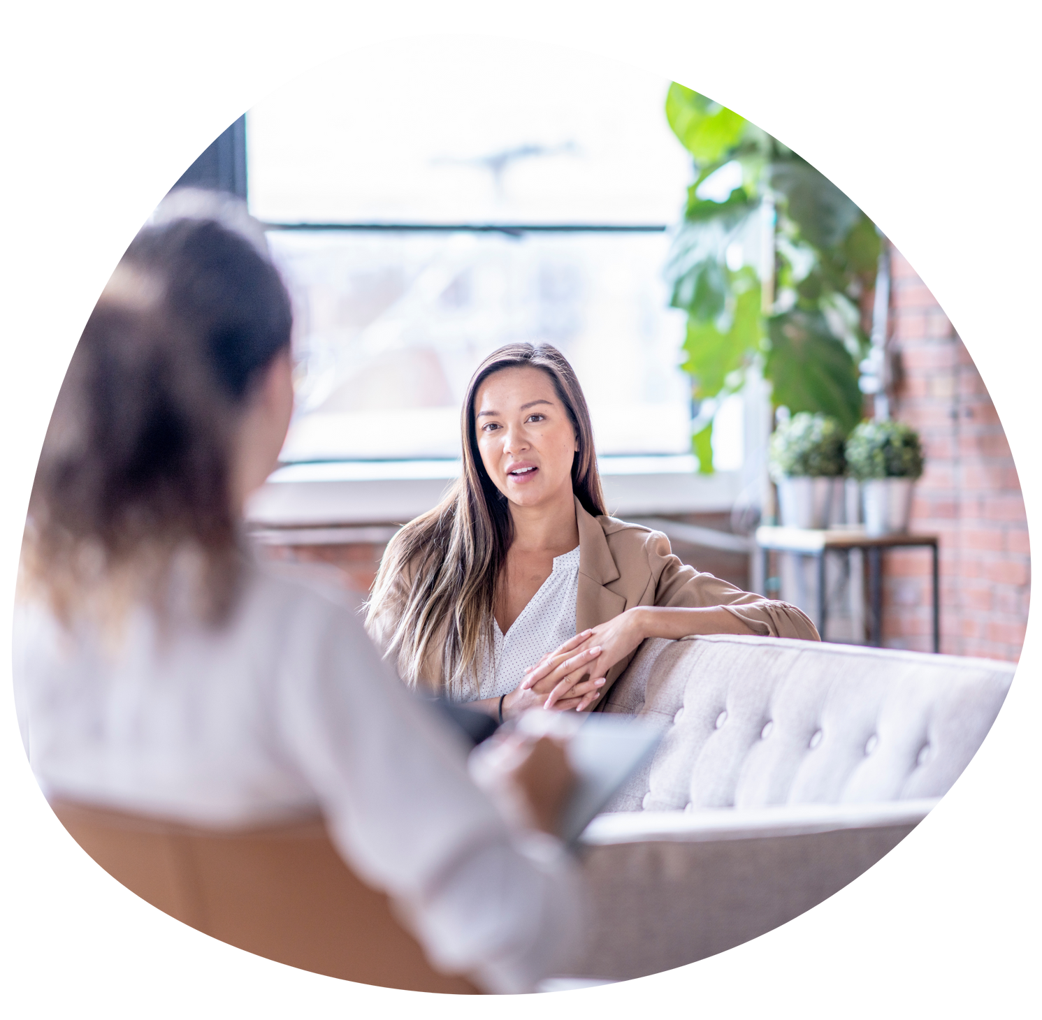 Two women talking in a bright living room, one sitting on a white sofa and smiling