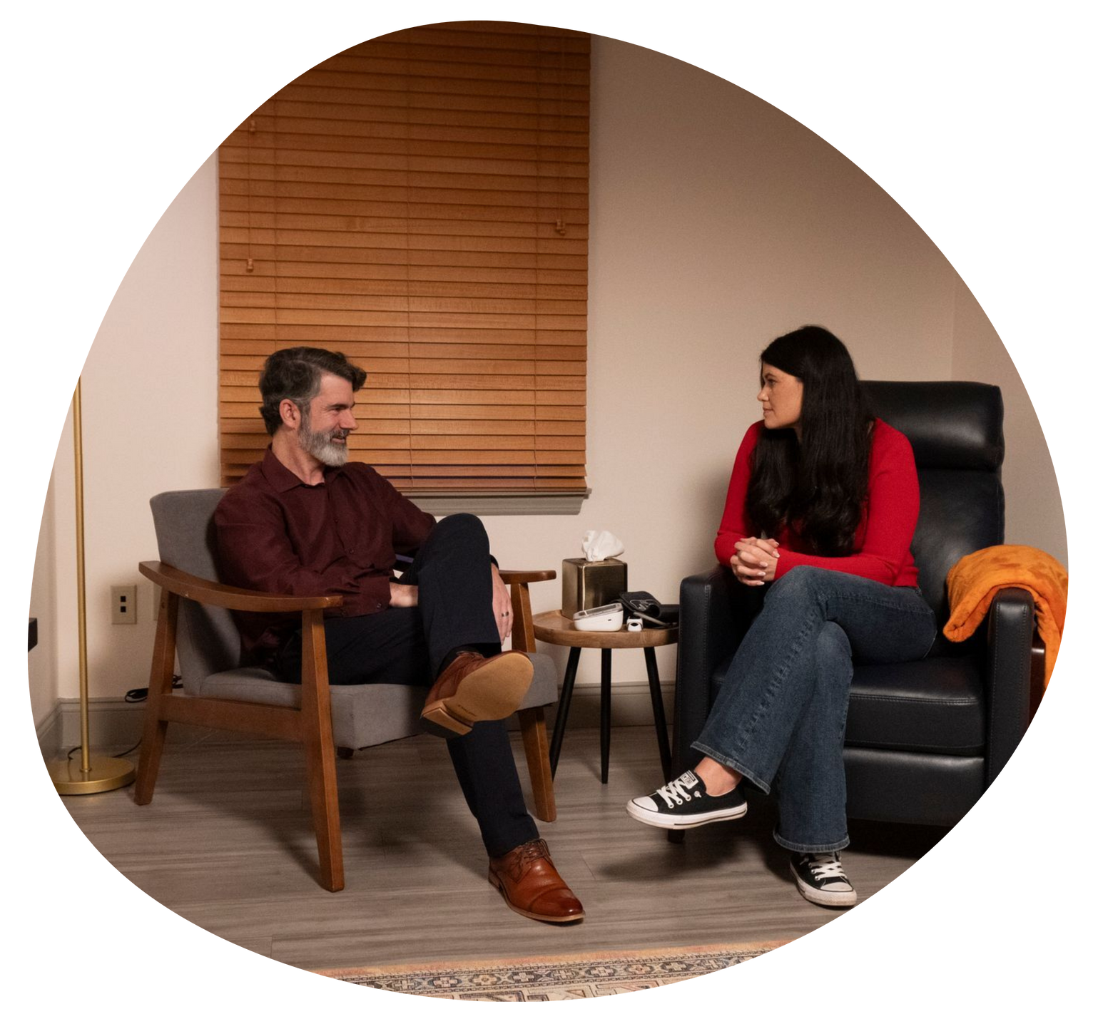 Two people talking in chairs in a cozy room with a window blind and small table between them