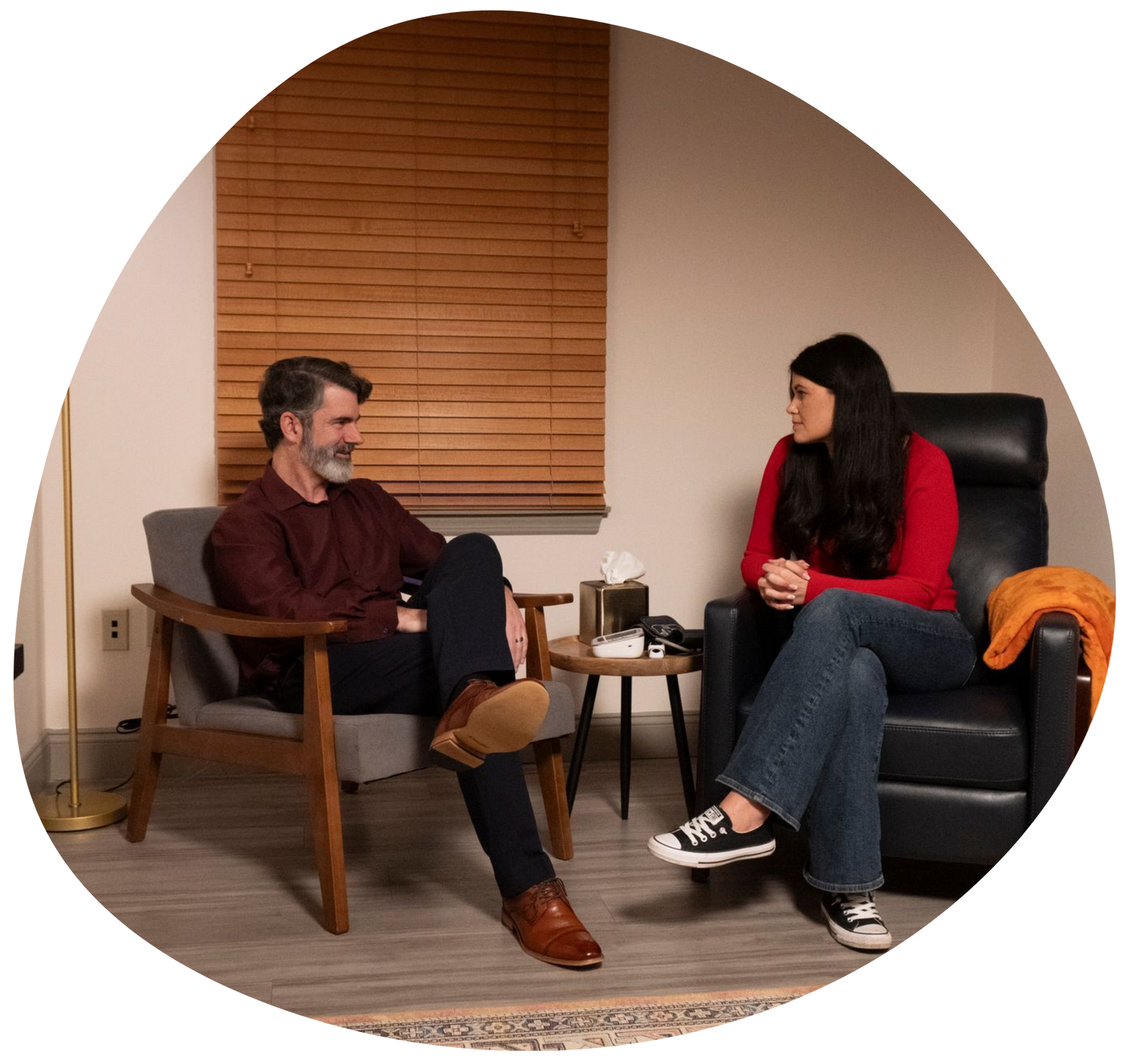 Two people seated in a living room, talking across small tables, with one in a red sweater and one in a maroon shirt.