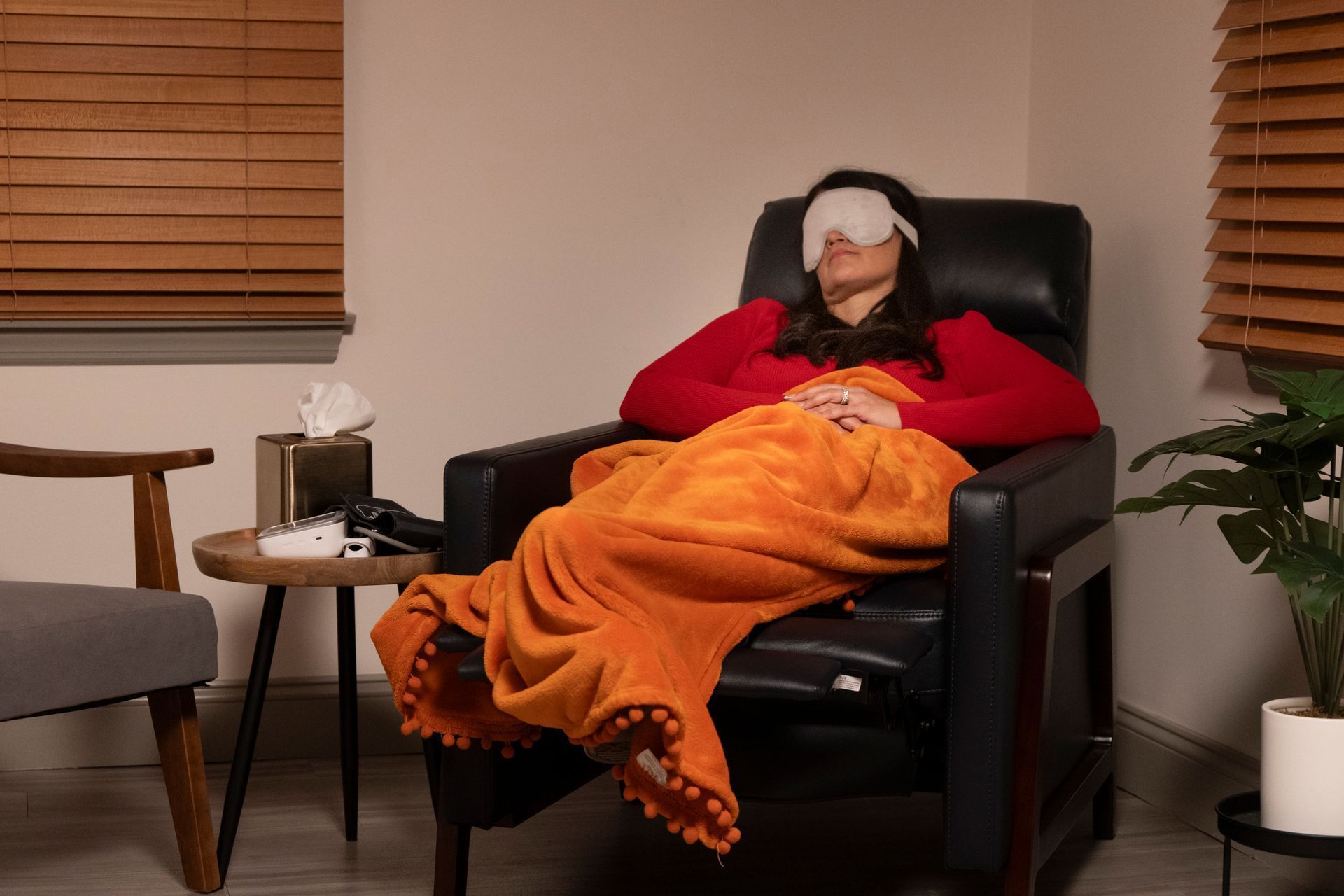 Person resting in a recliner under an orange blanket, wearing a sleep mask in a cozy living room.