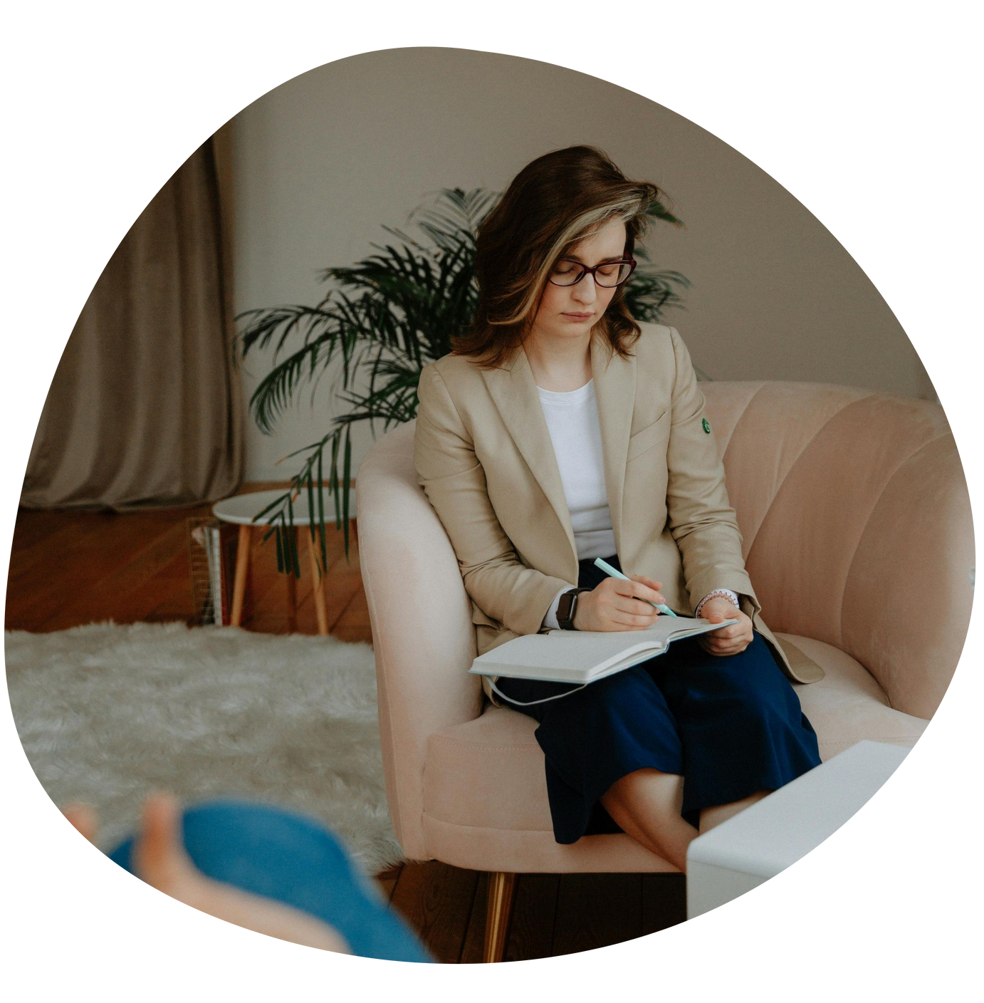 Woman in beige blazer taking notes in a notebook while seated in a cozy living room.