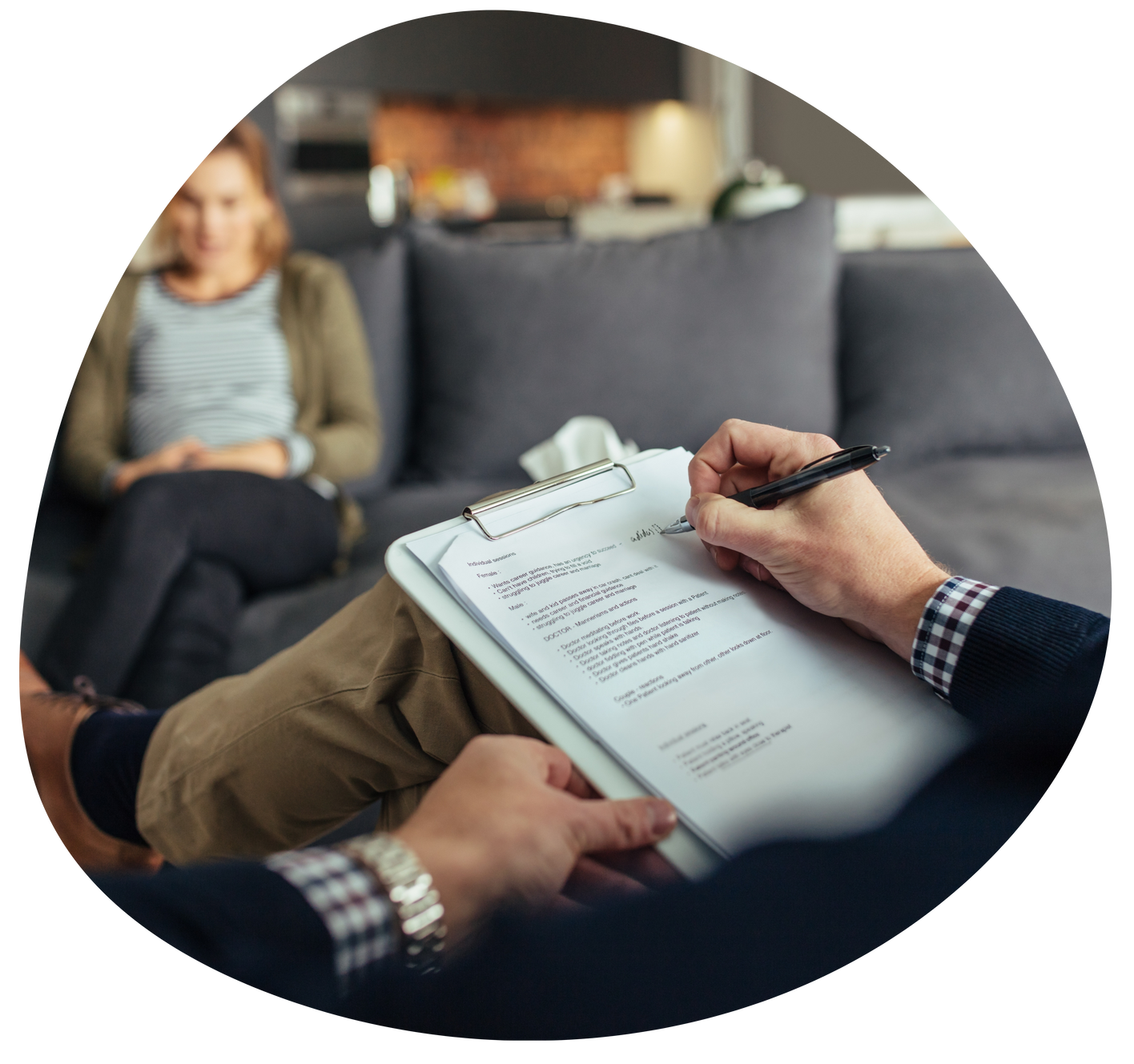 Therapist taking notes on a clipboard while speaking with a seated client on a couch