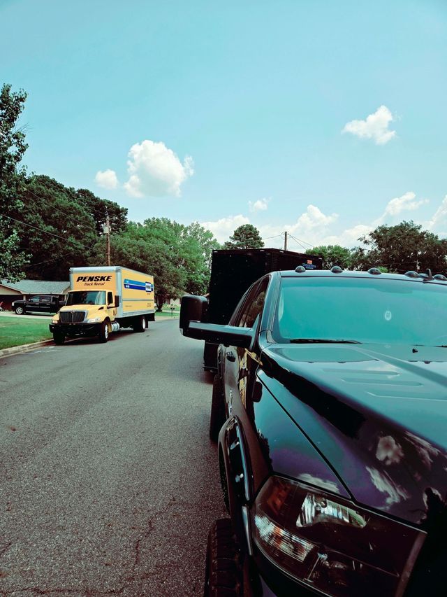 Black pickup truck on a road with a yellow truck ahead under a blue sky.