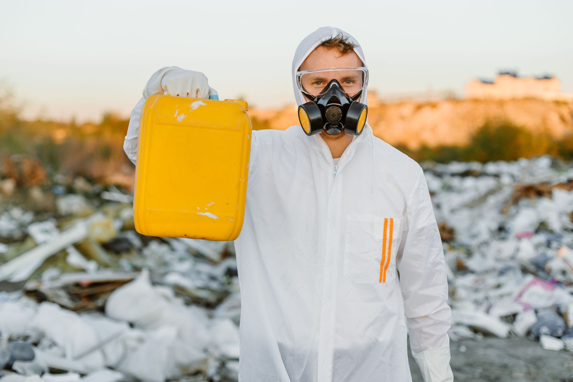 Person in white protective suit and mask holding a yellow container in a trash-strewn outdoor area.