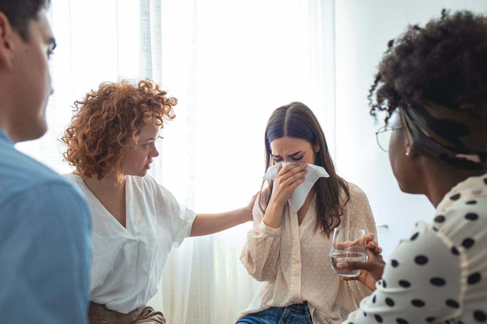 A woman in a support group receives comfort. A hand rests on her shoulder. Three others watch in a brightly lit room.