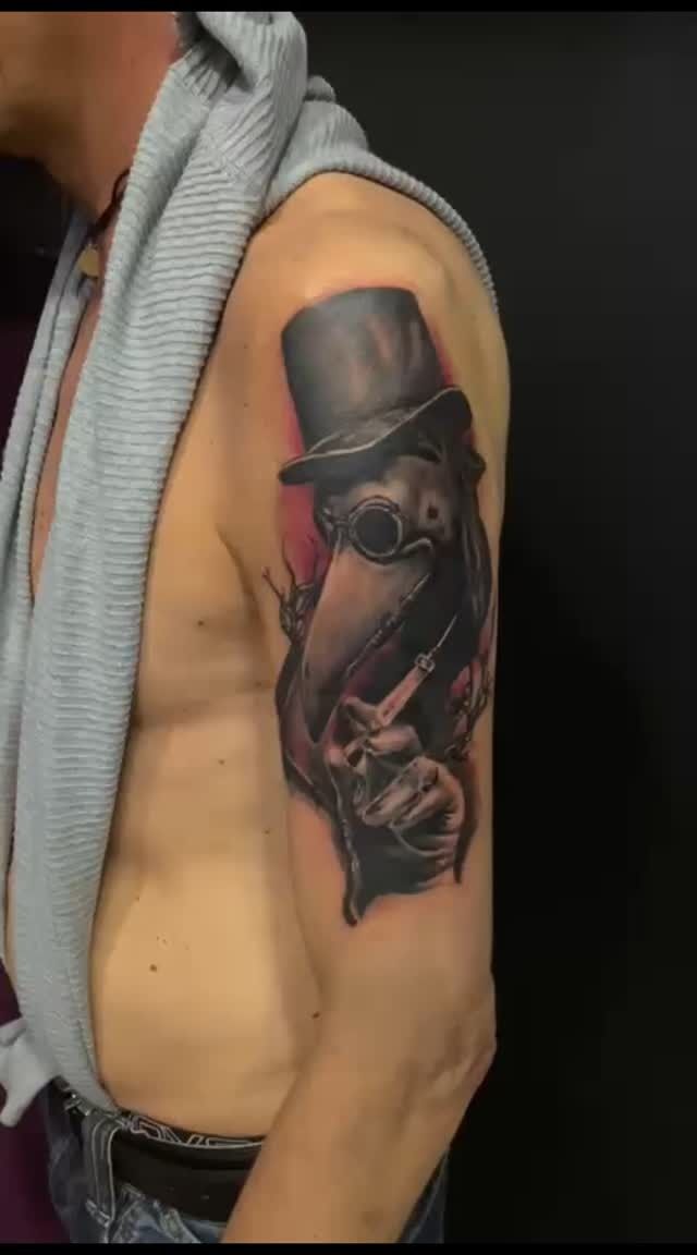 Tatuaggio di un medico della peste sulla parte superiore del braccio di un uomo. Il tatuaggio è in nero e grigio con sfondo rosso.