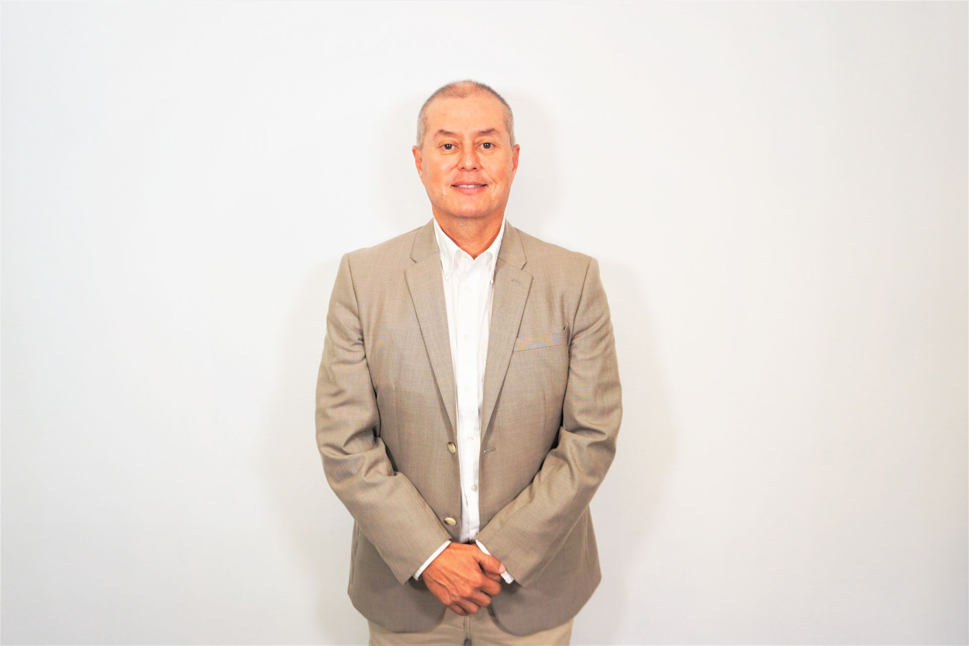 Man in a beige suit stands against a plain white background, hands clasped in front.
