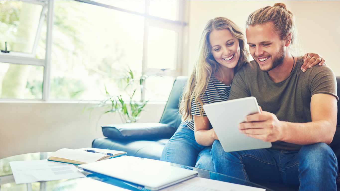 Couple smiling on a couch, looking at a tablet in a bright living room.