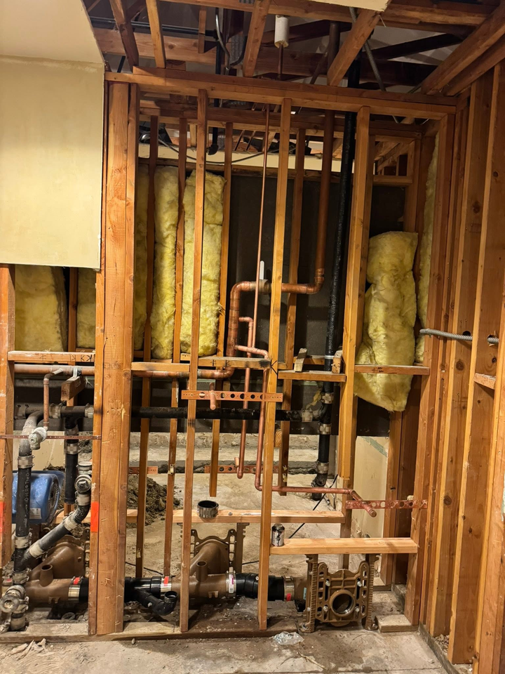 Exposed wood framing and pipes in a gutted room with debris on the floor and insulation in the walls