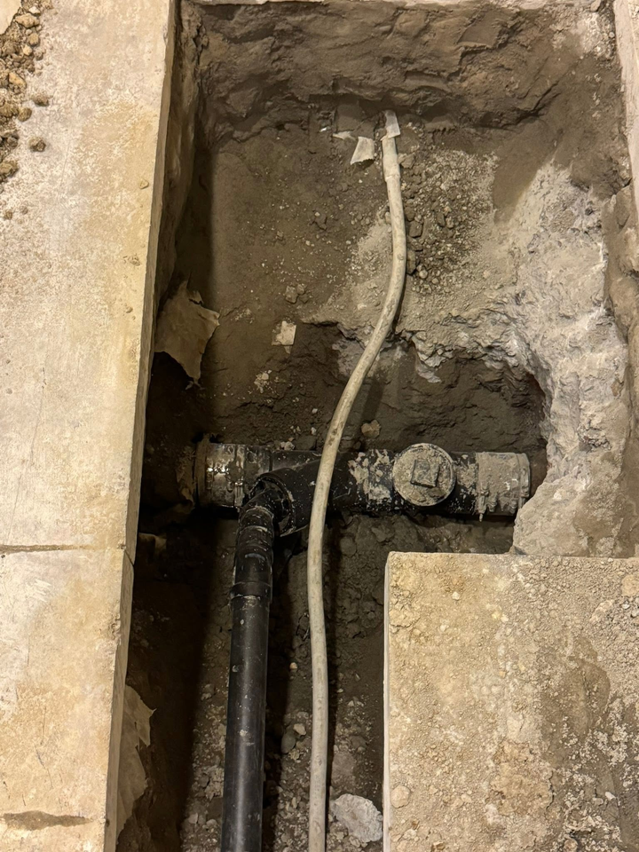 Open floor trench with exposed pipes and a white cable in rough concrete.