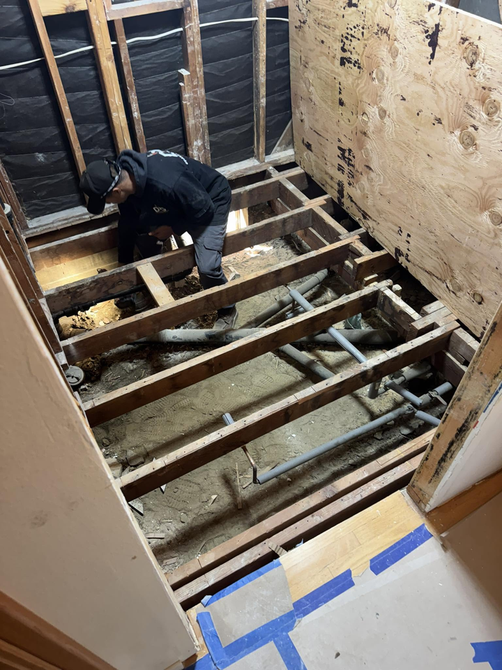 Worker in dark clothing repairing exposed floor joists in a stripped room with bare walls and blue painter’s tape.