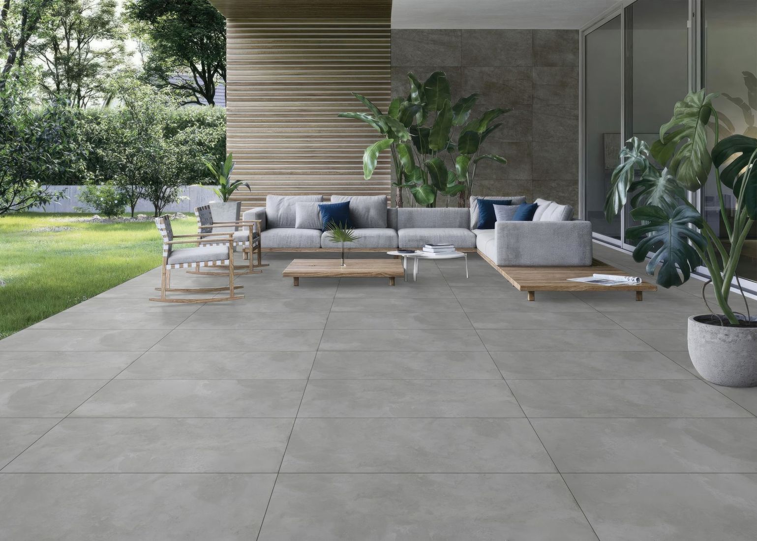 Modern outdoor patio with gray sectional seating, potted plants, and large stone pavers beside a lawn