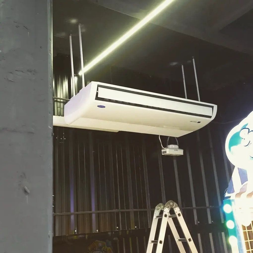 Unidad de aire acondicionado blanca montada en el techo, sobre cortinas negras y una escalera en un espacio interior.