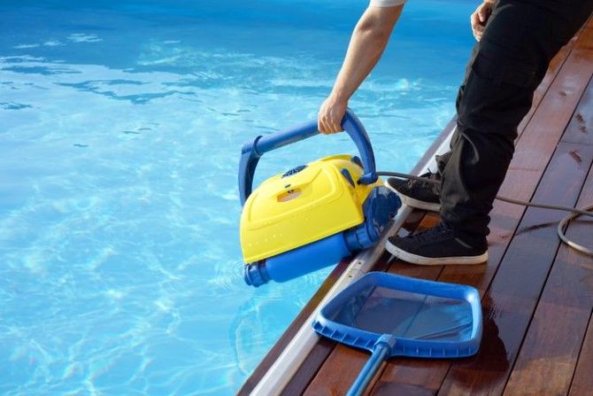 Person using a yellow pool vacuum beside a swimming pool, with a blue skimmer on the deck