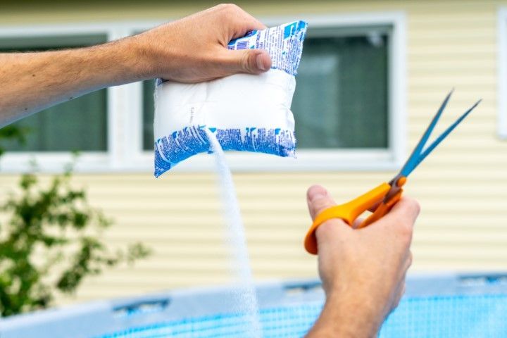 Hands holding a small bag of blue pool sanitizer pellets and orange scissors by a backyard pool