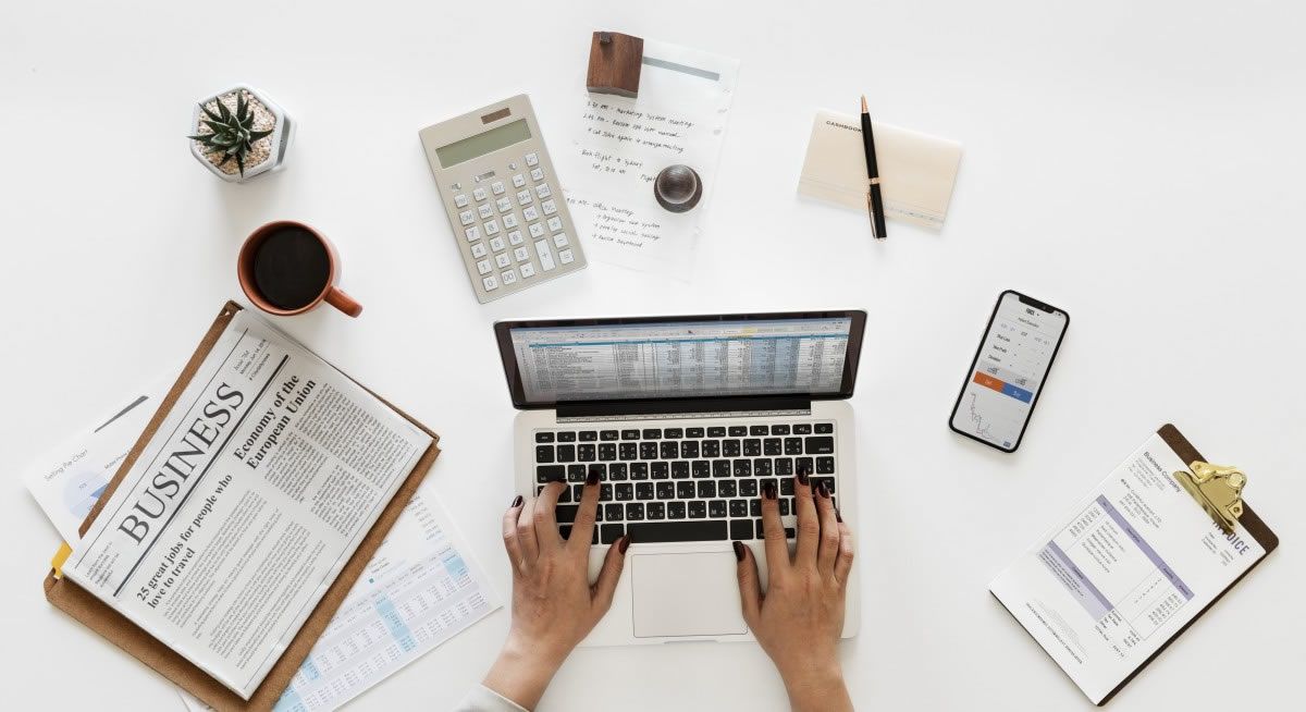 Manos tecleando en un portátil sobre un escritorio limpio con periódico, café, calculadora, teléfono y cuaderno.