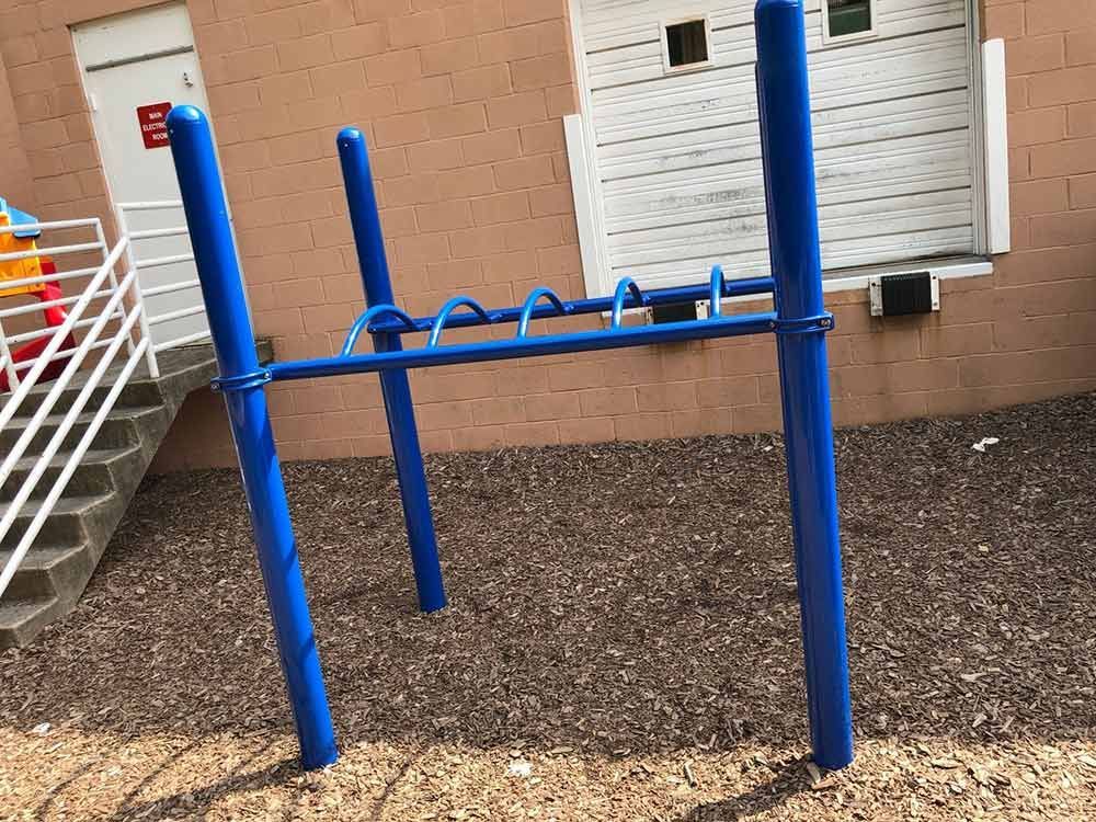 Blue outdoor exercise bars beside a building and stairs, over gravel ground.