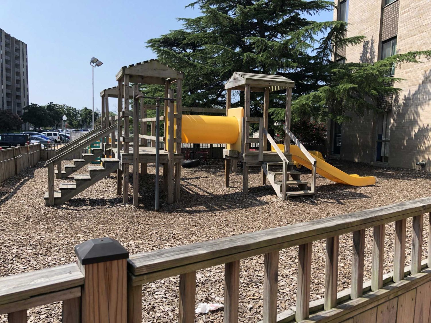 Outdoor playground with a yellow tunnel slide, climbing stairs, and wooden structures on a gravel surface.