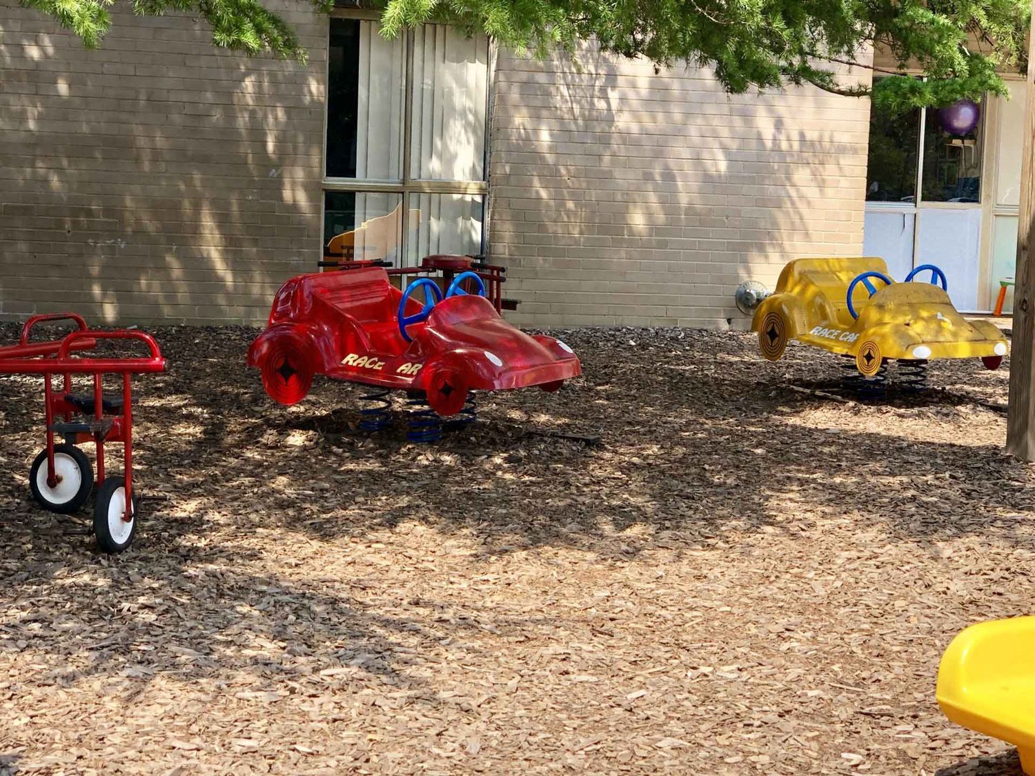 Red, yellow, and black toy cars parked on wood chips beside a building.