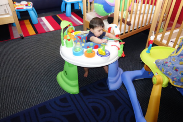 Baby at a colorful activity table in a playroom, surrounded by toys and play equipment.