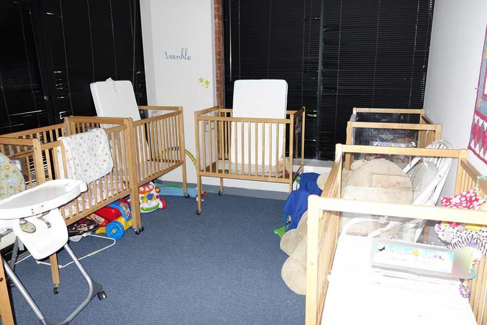 Empty daycare room with wooden cribs, white chairs, toys, and a blue carpet