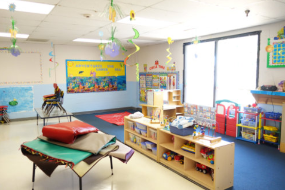 Bright classroom with shelves, colorful posters, hanging decorations, rugs, and a large window.