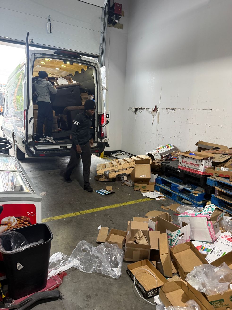 Person loading boxes into a truck inside a warehouse with scattered cardboard and pallets on the floor