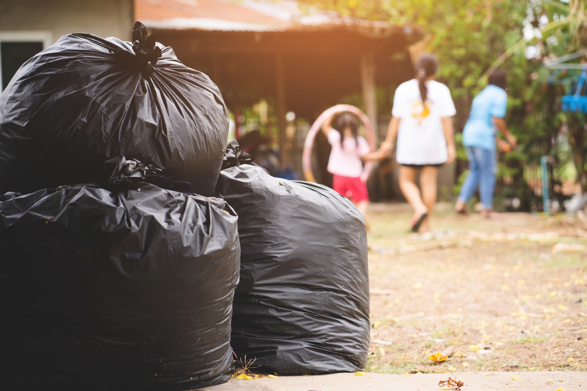Black garbage bags piled outdoors near a blurred path with people walking in the background