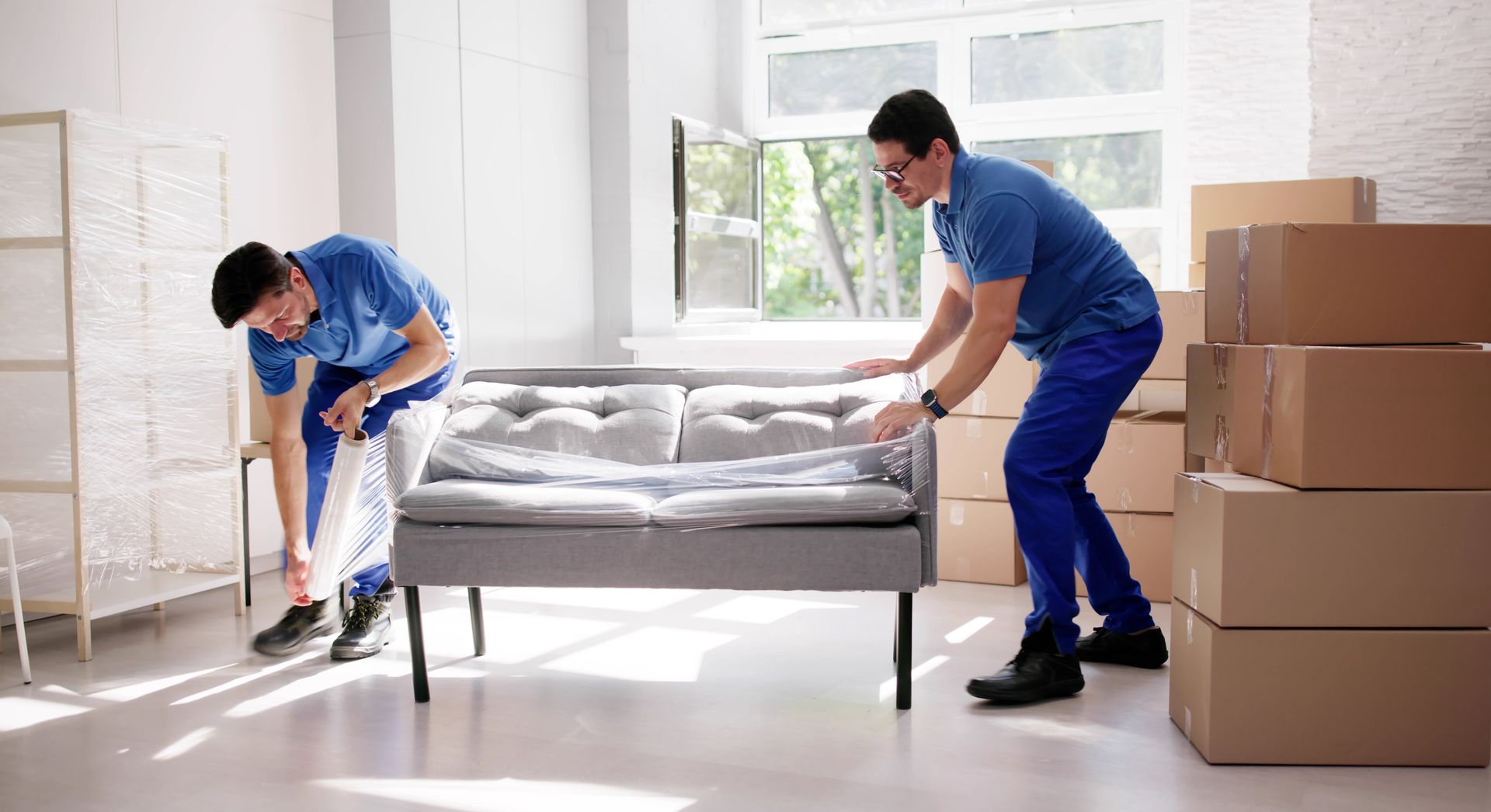 Two movers in blue uniforms carrying a gray sofa in a bright room beside stacked moving boxes