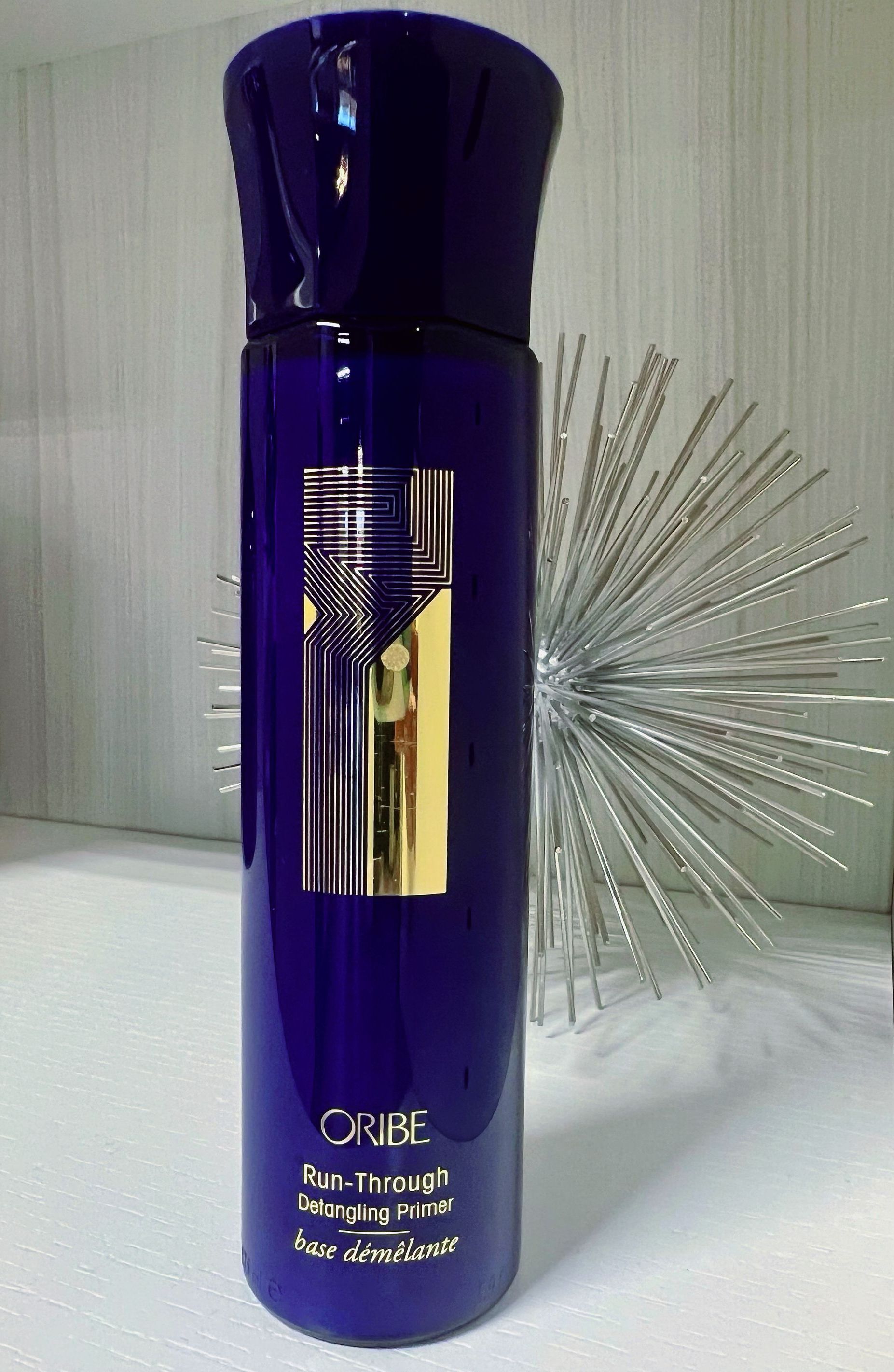 Tall dark blue bottle with shiny gold zigzag logo and Oribe Run-Through Detangling Primer printed at bottom.