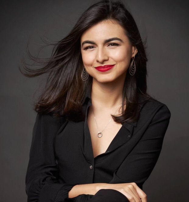 Young woman with medium skin tone and brunette shoulder length hair sitting smiling without showing teeth.