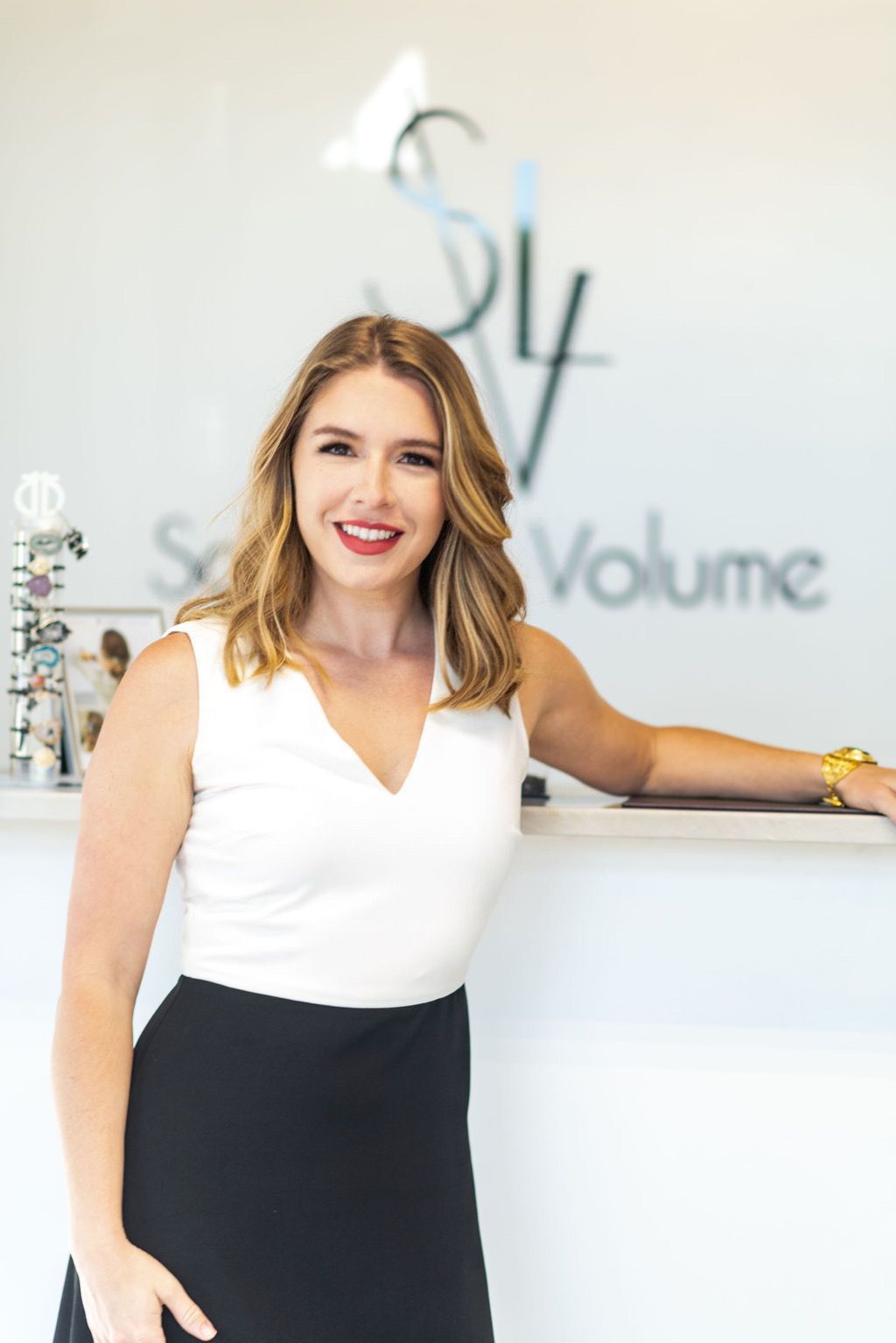 Smiling young woman with medium light skin tone and blonde hair stands in front of Salon Le Volume logo.